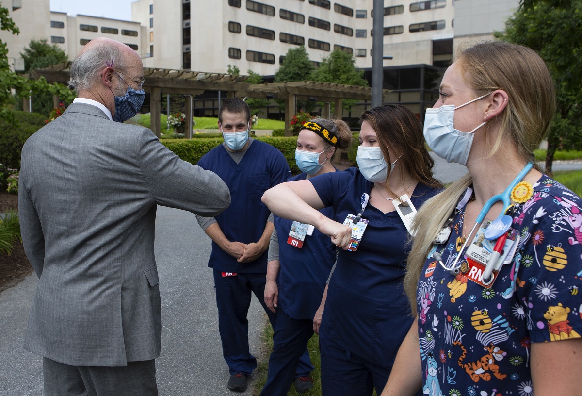 Gov. Tom Wolf, wearing a surgical mask and a suit, holds out an elbow to a woman in scrubs and a surgical mask, who offers her elbow in return. Three others in scrubs and surgical masks stand in a row. The widows and cement of Hershey Medical Center rise in the background.