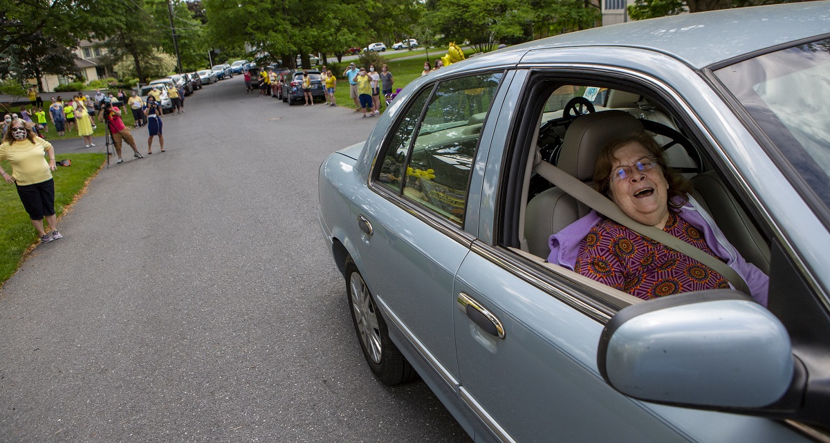 A woman in the passenger seat of a car smiles. Behind her, people line the roads and take photos.