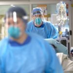 Two health care workers in personal protective equipment, including surgical masks and face shields wheel a gurney through a hospital hallway. Attached to the gurney are IV bags on a pole.