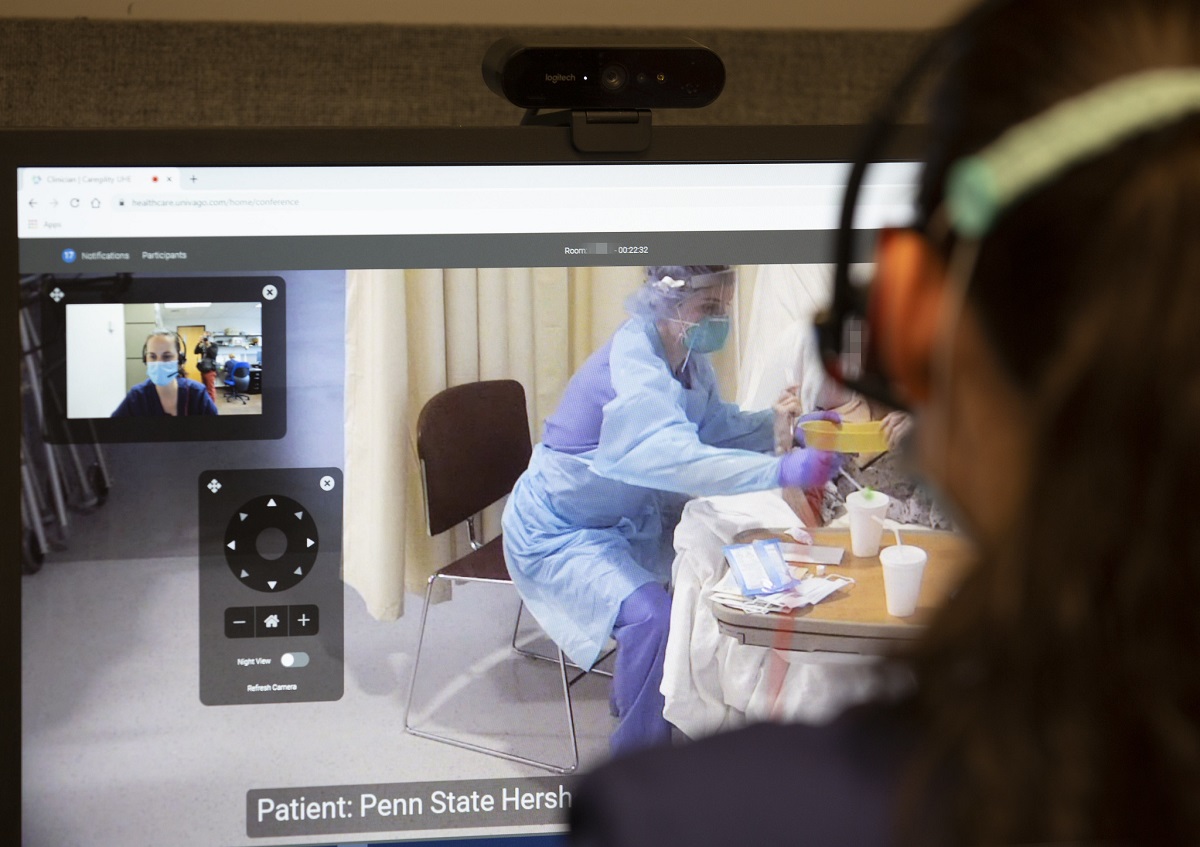 Image shows over the shoulder someone looking at a computer monitor. In one window on the monitor is the view of someone in a mask. To the right, someone in personal protective equipment works with a patient.