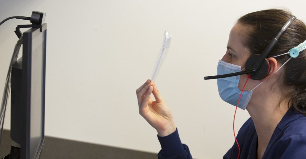 A person in profile wears a surgical mask and holds a toothbrush up to a computer monitor. The person holding the toothbrush also wears a headset. 