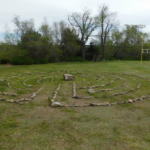 Rocks are laid out on the ground, forming an outer circle with paths inside it. The labyrinth is in a field, with a treeline visible behind it.