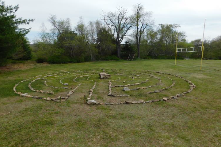 Rocks are laid out on the ground, forming an outer circle with paths inside it. The labyrinth is in a field, with a treeline visible behind it.