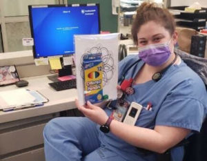 A woman wearing a medical face mask and scrubs sits at a desk, holding a packet with colored pencils and coloring sheets.