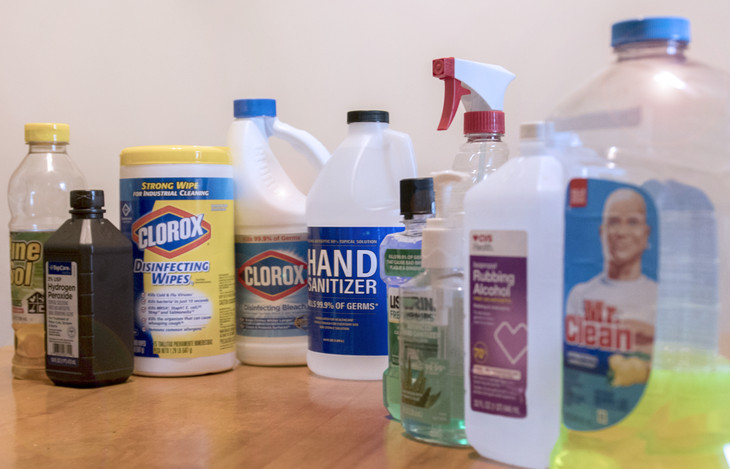 Ten household cleaners are pictured sitting on a table. They include Pine-Sol, hydrogen peroxide, Clorox disinfecting wipes, Clorox bleach, hand sanitizer, Listerine, rubbing alcohol, Mr. Clean and others.