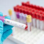 A stock photo shows a close-up view of a lab technician’s hand as it holds a test tube containing a substance that looks like blood and labeled HIV-positive.
