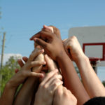 Close-up of several children extending their arms into the air to form a pyramid. In the background are trees, power lines and a basketball hoop.
