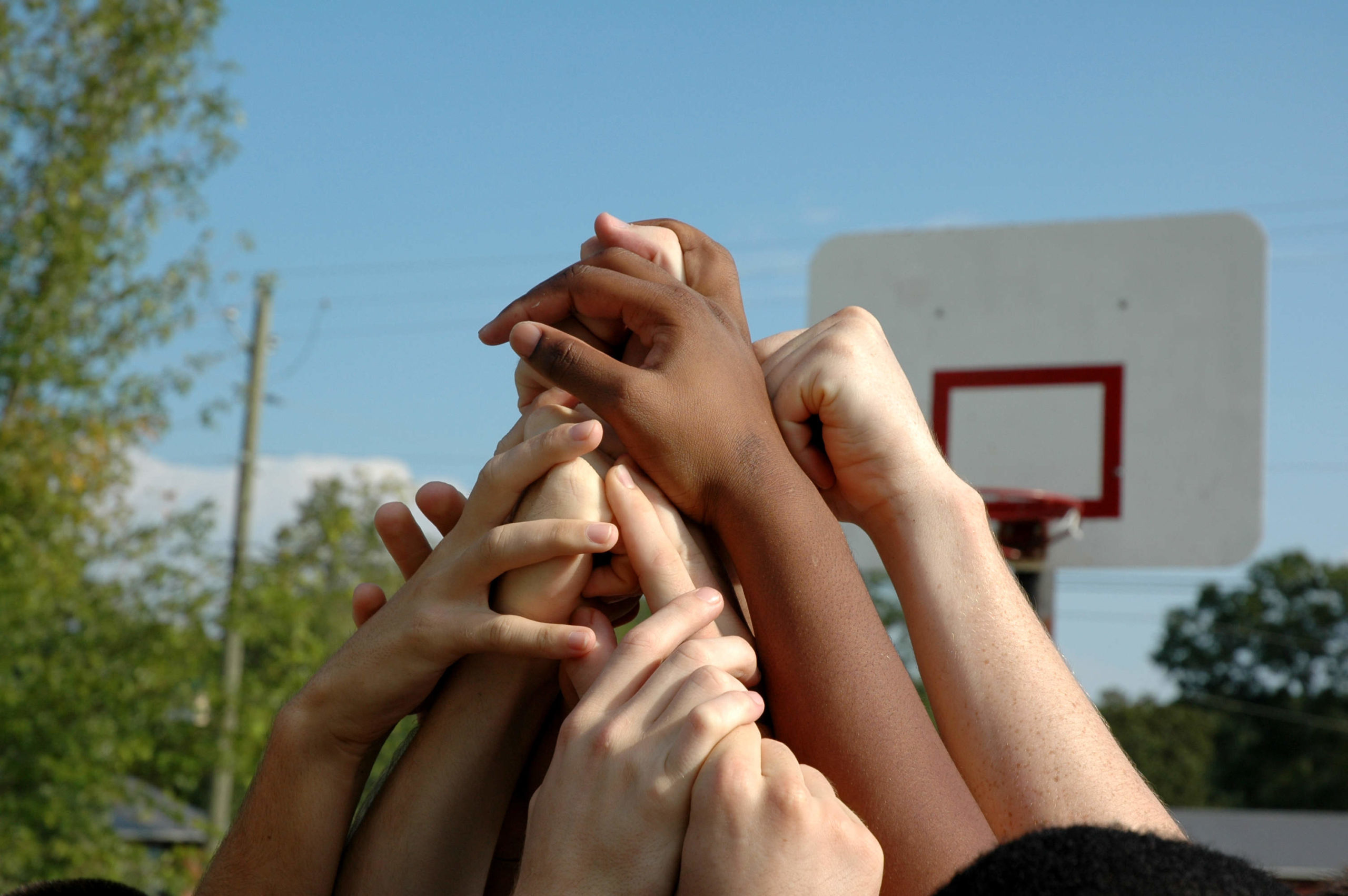 Close-up of several children extending their arms into the air to form a pyramid. In the background are trees, power lines and a basketball hoop.