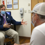 A male physician wearing a tie, Penn State Health coat, face mask and stethoscope, gestures with his hands while talking with a man seated across from him in a clinic room.