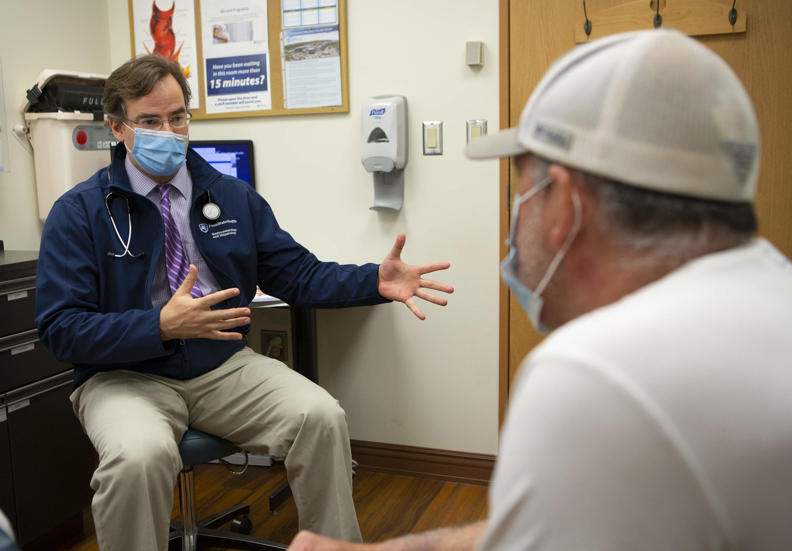 A male physician wearing a tie, Penn State Health coat, face mask and stethoscope, gestures with his hands while talking with a man seated across from him in a clinic room.