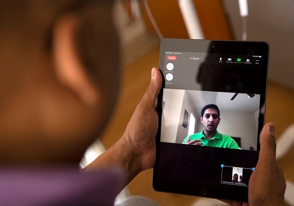 An image is taken from over the shoulder of ap erson. The person is holding an iPad, and the image of a student is on the screen.