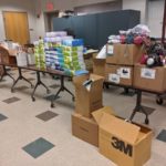 Boxes are seen sitting on a table, which is full. More boxes are on the floor. They are all filled with personal protective equipment.