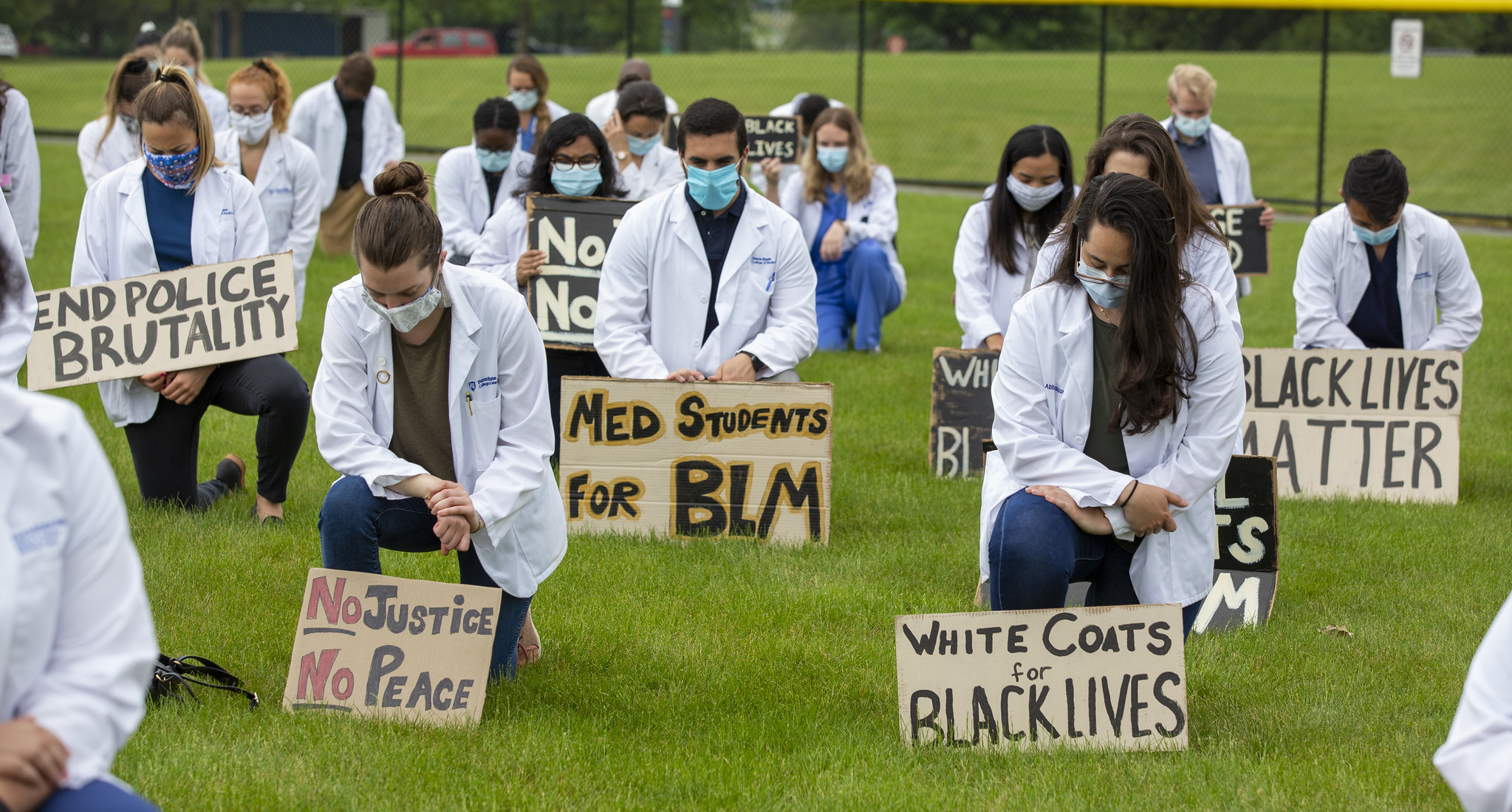 A group of people are seen spaced apart, kneeling on a green lawn. They hold signs with statements such as White Coats for Black Lives, No Justice No Peace, End Police Brutality, Black Lives Matter and Med Students for BLM.