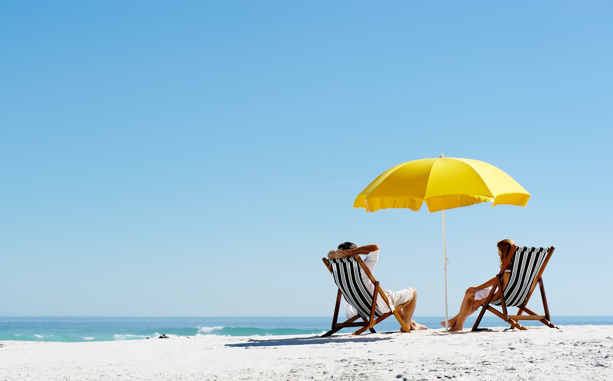 Two people sit in chairs on the beach under an umbrella.