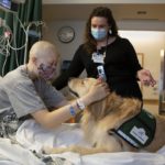 Emma Munger wears a mask and leans forward in a hospital bed to pet Becky, a golden retriever, who stares into Munger’s eyes. Behind them stands a woman in a mask and lanyards.