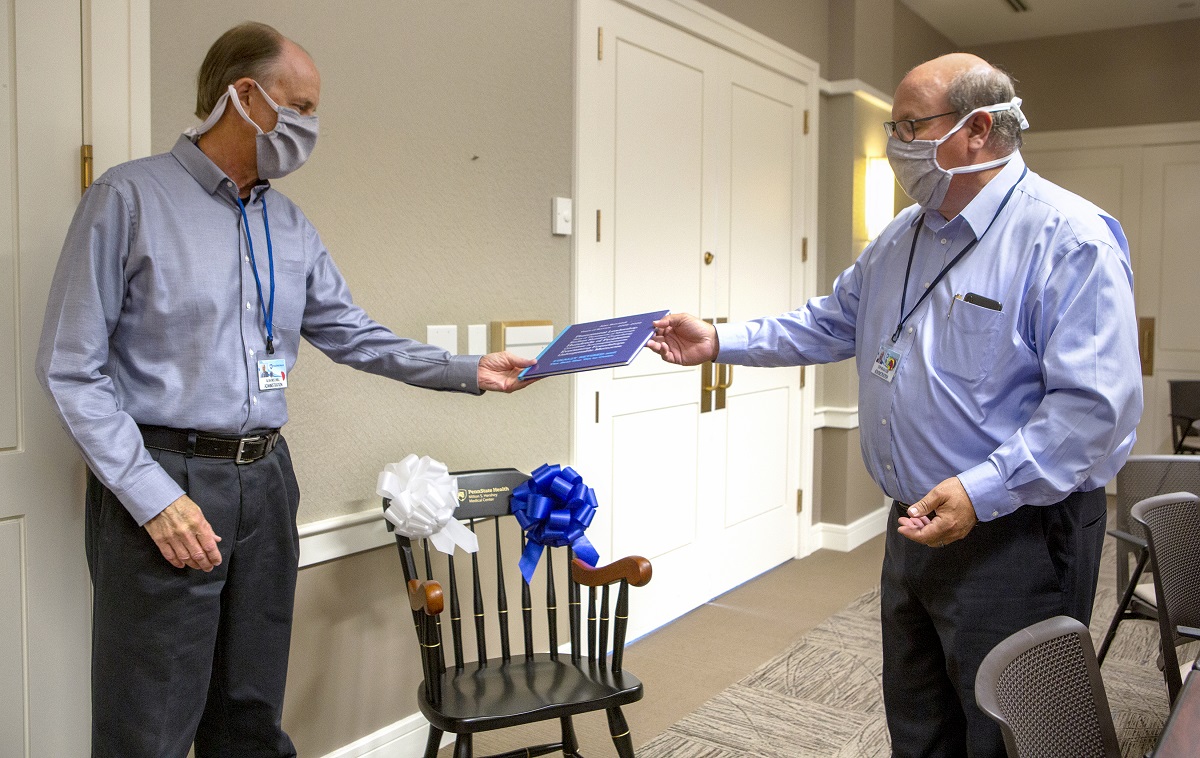 Penn State Health CEO Steve Massini hands a memory book to Alan Brechbill. Both men are wearing dress shirts, trousers and masks. A chair with a Penn State Health Milton S. Hershey Medical Center logo and two bows on it is in the background.