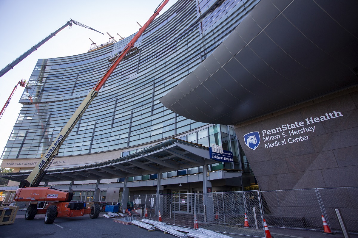 Two cranes sit in front of the entrance to the Children’s Hospital. A chain link fence wraps around the front of the building. Construction cones and other items are scattered about.