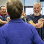 Hank Shipman smiles broadly as he stretches his arms out in front of himself and bends down his fingers. He wears a T-shirt and has a beard and moustache. Other rehab patients smile and laugh in the background as they do similar stretches. The instructor is seen from behind.