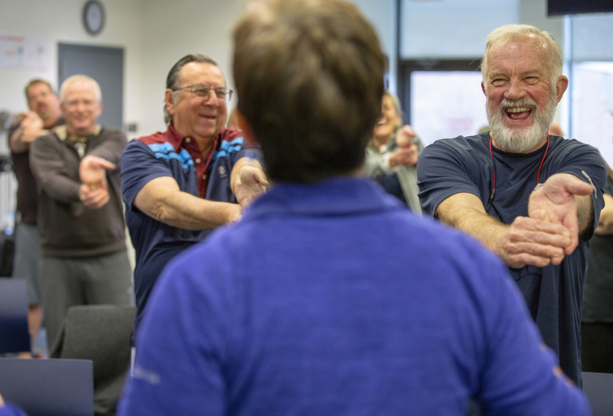 Hank Shipman smiles broadly as he stretches his arms out in front of himself and bends down his fingers. He wears a T-shirt and has a beard and moustache. Other rehab patients smile and laugh in the background as they do similar stretches. The instructor is seen from behind.