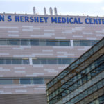 A building with the words "Milton S. Hershey Medical Center" on it rises through the center of the photo. A portion of a closer building with several windows is at right.