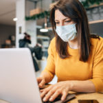 A woman in a face mask is sitting at a desk using a laptop.
