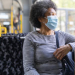 A woman sits on a bus, with her left arm resting on the window rail. She looks out the window. She's wearing a surgical mask.