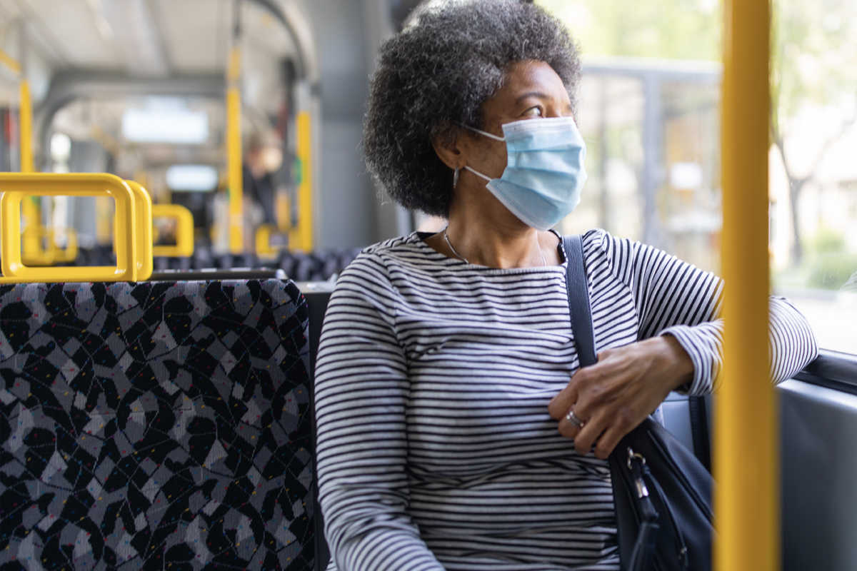 A woman sits on a bus, with her left arm resting on the window rail. She looks out the window. She's wearing a surgical mask.