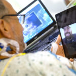 A man in a hospital gown in his hospital bed holds up an iPad on which a man is smiling back at him. A laptop computer is in the background, slightly out of focus.