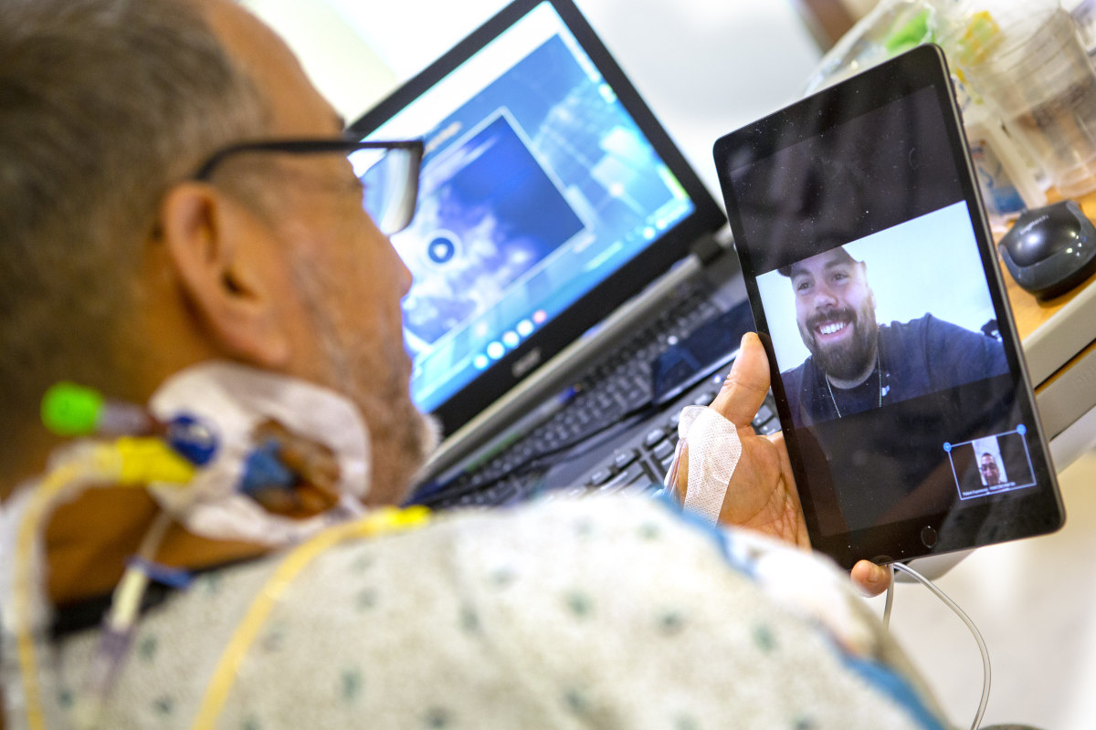 A man in a hospital gown in his hospital bed holds up an iPad on which a man is smiling back at him. A laptop computer is in the background, slightly out of focus.