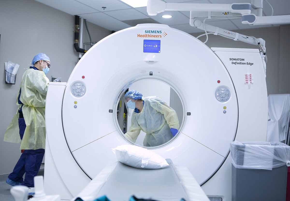 Un hombre y una mujer con máscaras faciales, guantes y fundas desechables para el cuerpo y la cabeza desinfectan una sala de tomografía computarizada después de ser utilizada por un paciente COVID-19 en el Centro Médico St. Joseph.