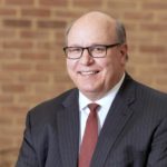 Steve Massini, Penn State Health CEO, poses in front of a brick wall. He is wearing glasses and a suit and tie. His hands are folded in front of him, and he is wearing a watch and wedding ring.