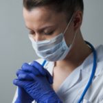 A female doctor wearing a white coat, face mask and gloves, bows her head and closes her eyes. Her hands are raised and her fingers are interlocked. A stethoscope is wrapped around her neck.