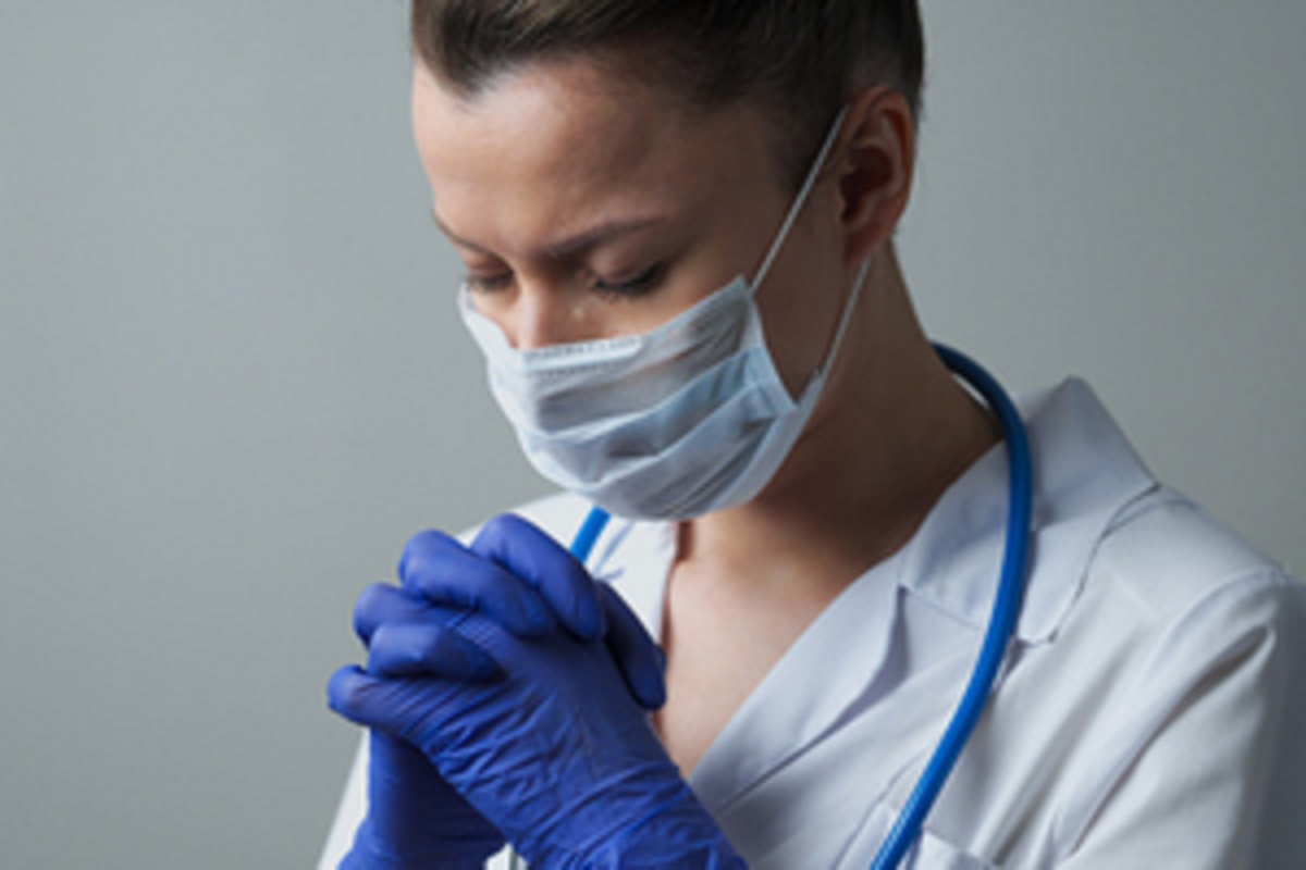 A female doctor wearing a white coat, face mask and gloves, bows her head and closes her eyes. Her hands are raised and her fingers are interlocked. A stethoscope is wrapped around her neck.