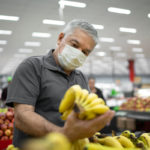 A man stands in the produce section of a grocery store, holding up a bunch of bananas and looking at them. He is wearing a face mask. Other shoppers are in the background.