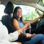 A woman sitting in the passenger seat of a car hands a key to a teenage girl sitting in the driver’s seat. Both look at each other, smiling.