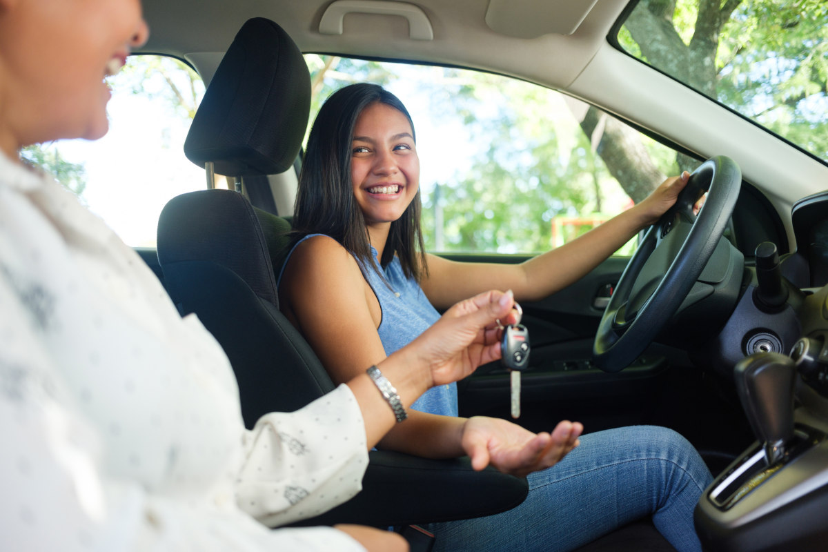 A woman sitting in the passenger seat of a car hands a key to a teenage girl sitting in the driver’s seat. Both look at each other, smiling.