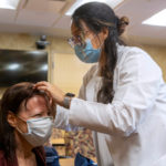 A woman in a white physician's coat looks at the top of a patient's head