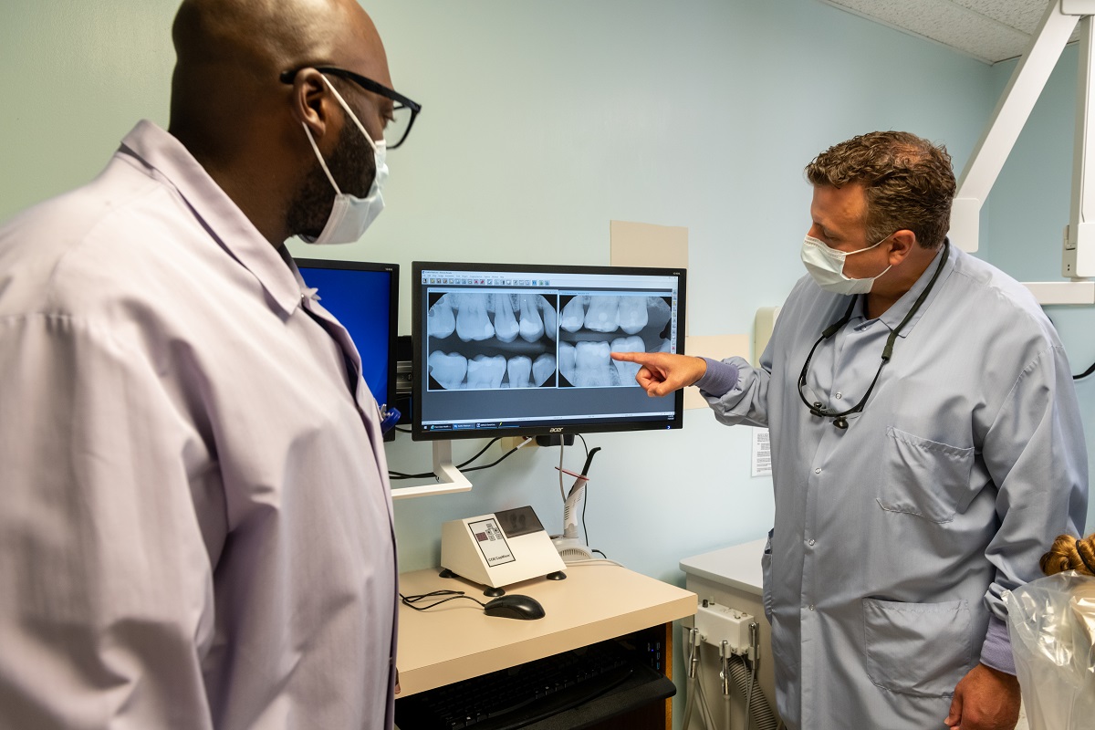 Dr. Larry Trubilla, wearing a lab coat and glasses hanging from his neck, points at the illuminated X-Ray image of a set of teeth. Dental resident Jowhar Brown, wearing glasses and a surgical mask, looks on.