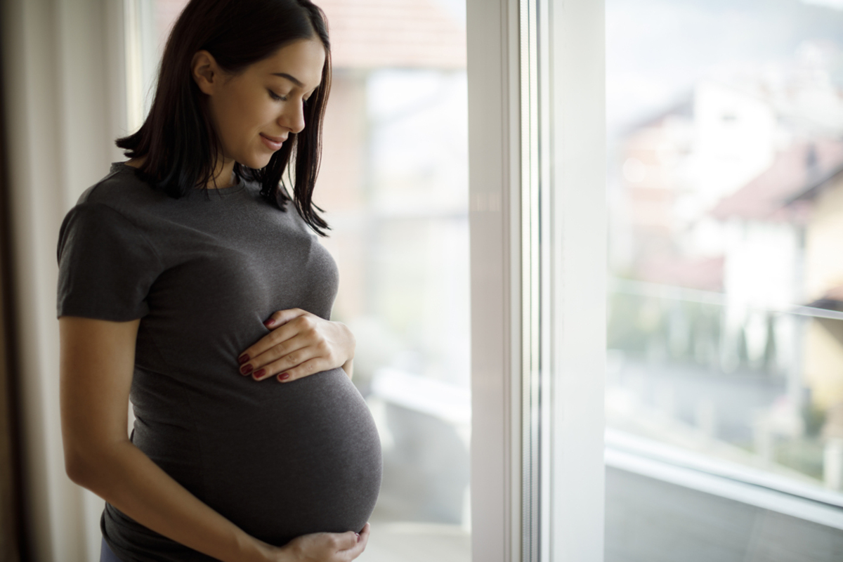 A pregnant young woman with shoulder-length hair puts her hands on her belly. She is standing next to a window.