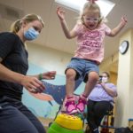 Joella Migliori jumps over a plastic stepping stone in a physical therapy room. Physical therapist Emily Hoffman, who has her hair in a pony tail and wears a face mask, kneels beside her. Joella, who wears glasses and has short hair pulled back in a barrette, wears a T-shirt that says “Spoiled by Daddy, loved by Mommy” with shorts and sneakers. Janette Migliori, Joella’s mother, sits in a chair in the background and holds up to phone to take a picture.