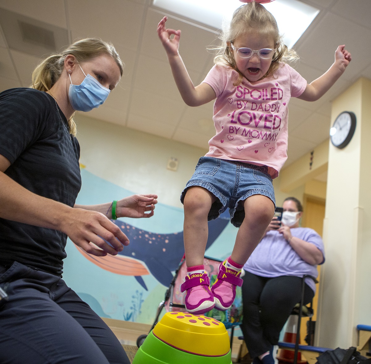 Joella Migliori jumps over a plastic stepping stone in a physical therapy room. Physical therapist Emily Hoffman, who has her hair in a pony tail and wears a face mask, kneels beside her. Joella, who wears glasses and has short hair pulled back in a barrette, wears a T-shirt that says “Spoiled by Daddy, loved by Mommy” with shorts and sneakers. Janette Migliori, Joella’s mother, sits in a chair in the background and holds up to phone to take a picture.