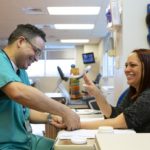 St. Joseph occupational therapist Julio Torres, left, smiles as he touches the hand of patient Diana Espinosa. Torres is wearing scrubs and glasses. Espinosa has long hair and is wearing a top with lacy sleeves. Behind them are balls hanging on the wall and a therapy bike.