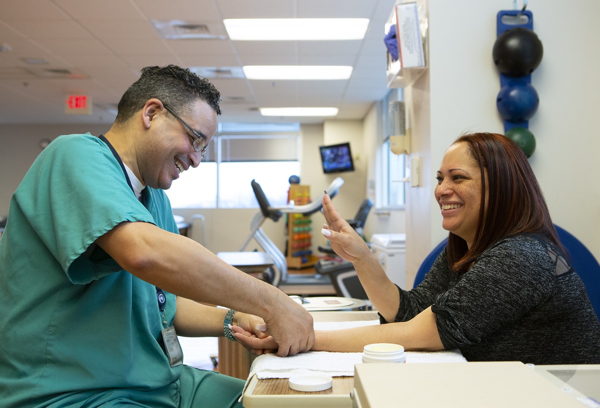 St. Joseph occupational therapist Julio Torres, left, smiles as he touches the hand of patient Diana Espinosa. Torres is wearing scrubs and glasses. Espinosa has long hair and is wearing a top with lacy sleeves. Behind them are balls hanging on the wall and a therapy bike.