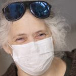 An older woman smiles behind a face mask. Her sunglasses rest on top of her head.