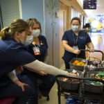 A woman in a surgical mask pushes a cart full of fruit and other snacks. Two women in scrubs and surgical masks pick from the items.