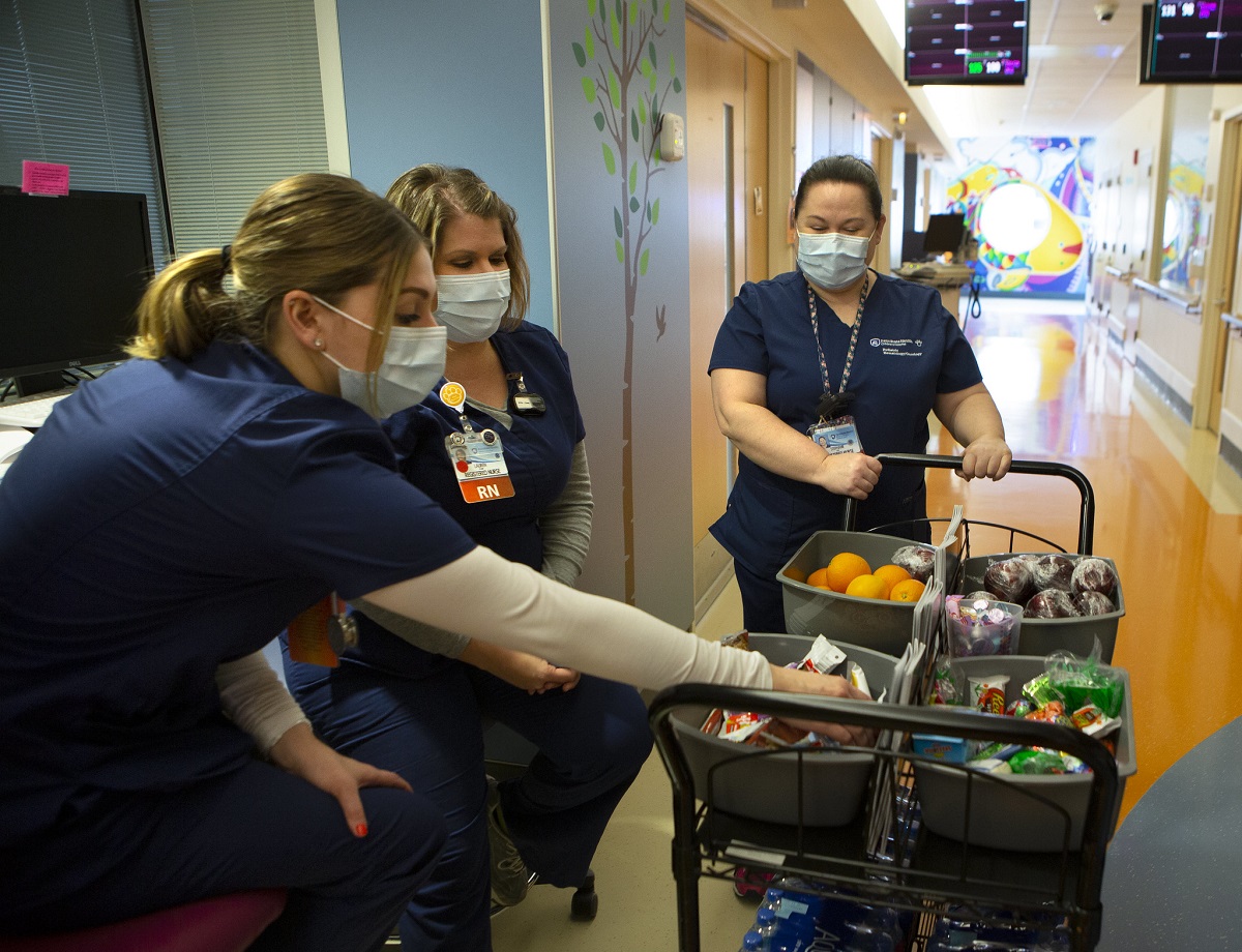 A woman in a surgical mask pushes a cart full of fruit and other snacks. Two women in scrubs and surgical masks pick from the items.