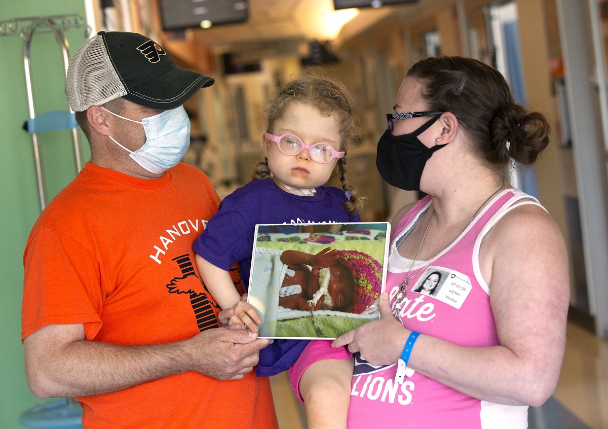 Jon Beckner, wearing a baseball cap and a T shirt, hold a little girl in pigtails and glasses. His wife holds the little girl from the other side. They also hold two sides of a photo of a premature baby with in a knitted cap.