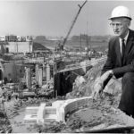 An older gentleman wearing a suit and tie, eyeglasses and a hard hat poses in front of the Hershey Medical Center construction site.