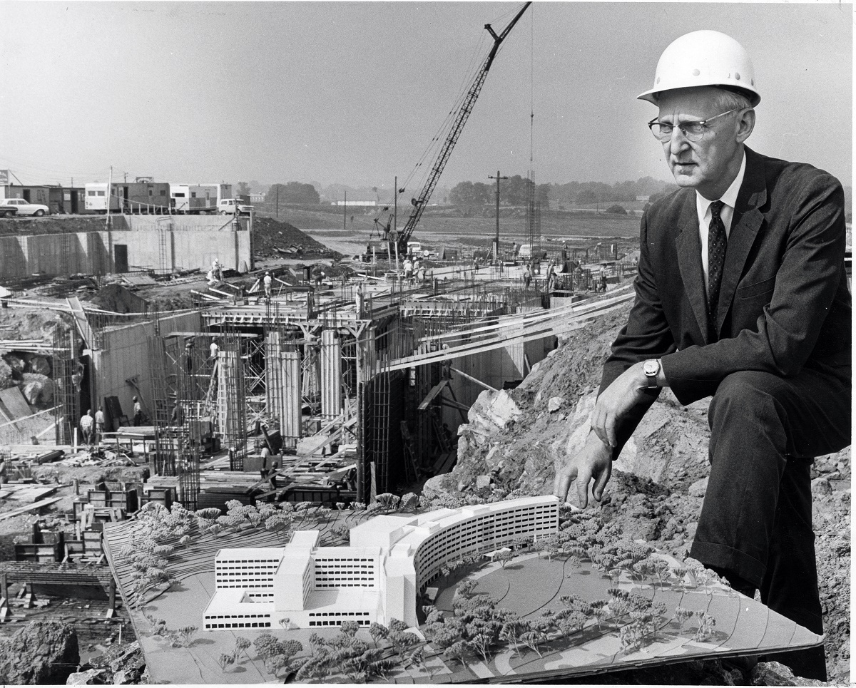 An older gentleman wearing a suit and tie, eyeglasses and a hard hat poses in front of the Hershey Medical Center construction site.