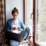A young woman wearing glasses sits by a window looking at a cell phone in her hands. A laptop rests on her leg.